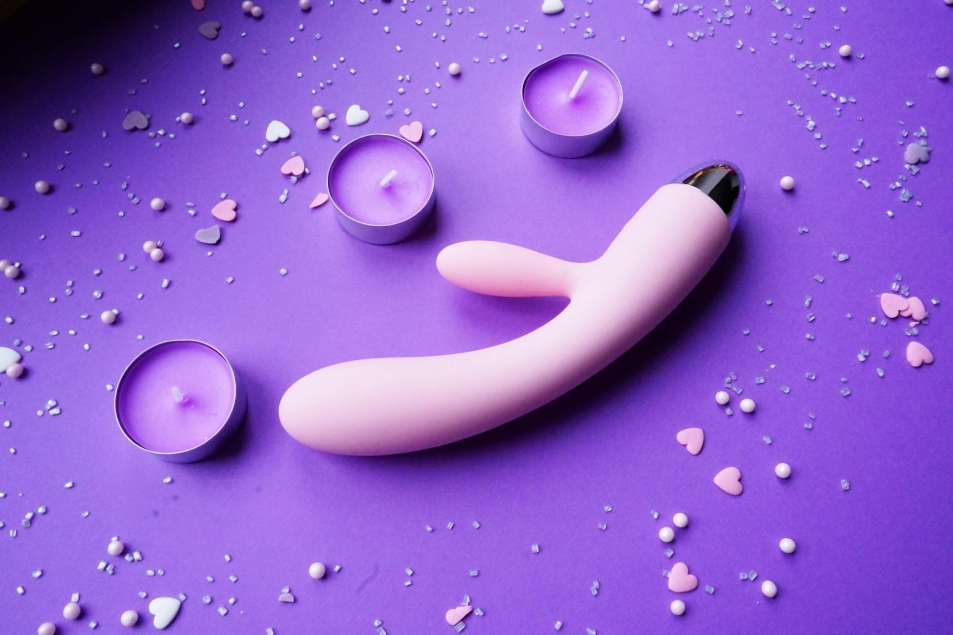A pink vibrator displayed on a purple background with heart confetti and candles, showing a romantic couples setting.